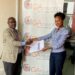 Gordon Asimwe, Marketing Manager GA, Insurance handin over the travel documents to Emily Mahero, the Mayhem Winner at GA Offices.